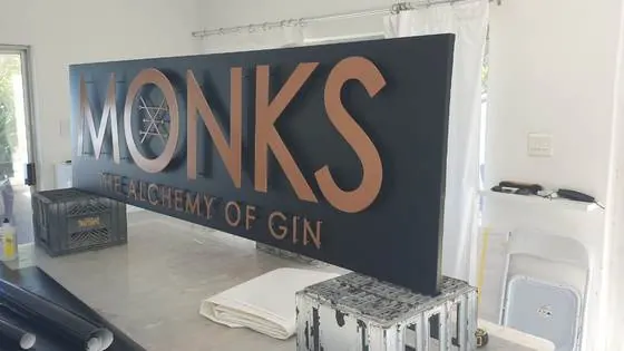 Monks gin sign with flat-cut copper and brushed metal lettering on a dark panel
